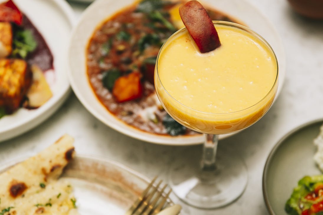 A mango lassi cocktail on a table surrounded by Indian share dishes
